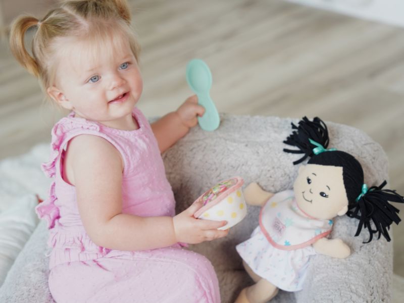 Toddler age girl seated in a gray child-sized chair holding a pretend food bowl and spoon, pretending to feed her soft doll that is also seated in the chair wearing a fabric bib.