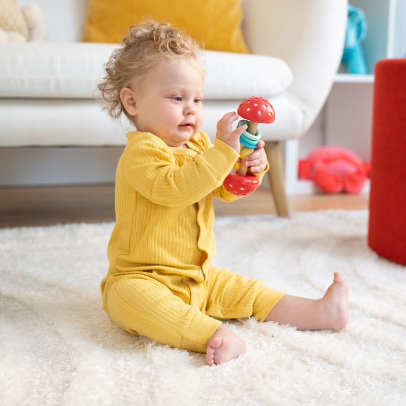 Baby girl seated in room scene holding a red, blue and natural wood colored barbel rattle. Girl is looking at the toy in her hands while in a living room scene.