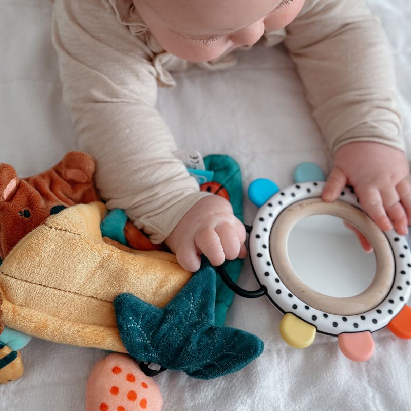 Baby laying on tummy reaching for a round mirror grasping toy with colorful teething nubs around the edge and a soft book laid nearby.