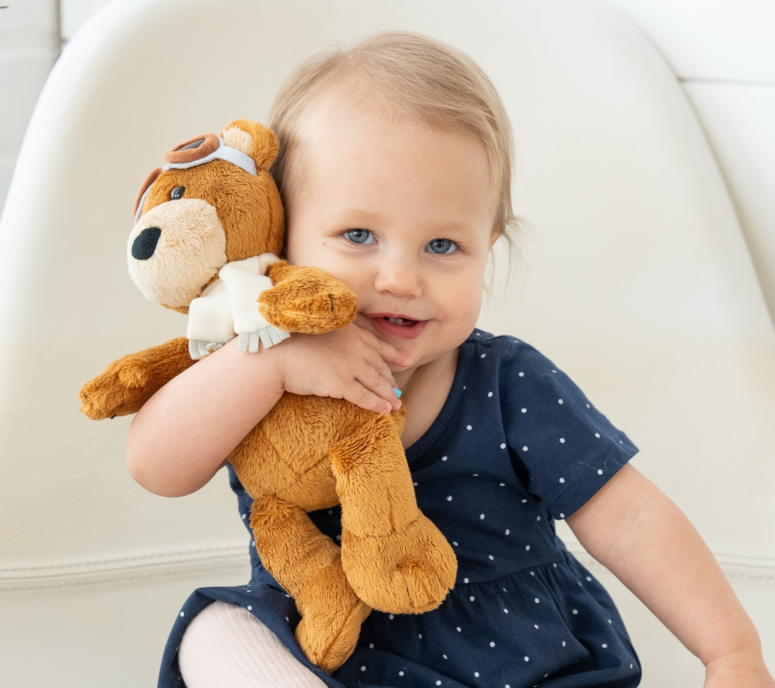 Smiling toddler hugging a plush aviator bear while sitting on a white chair.