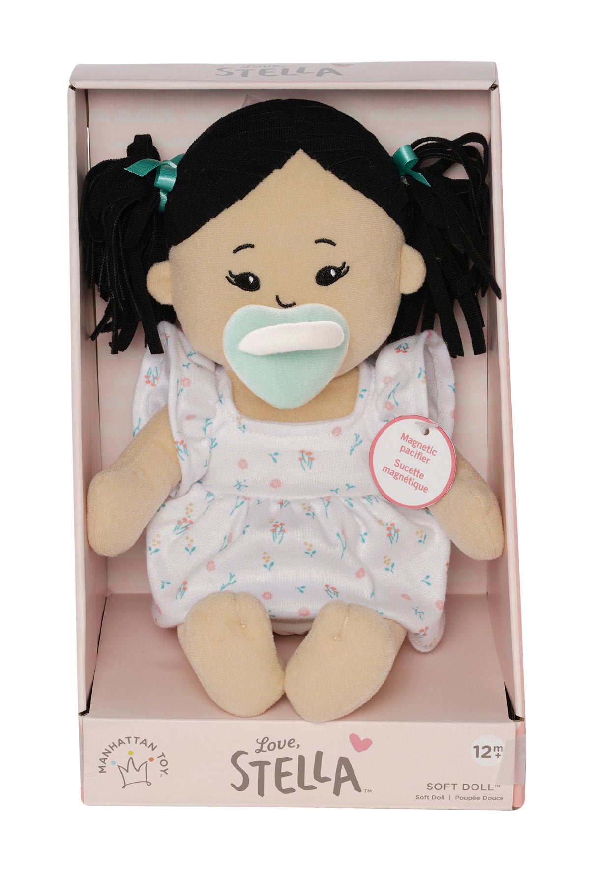 Soft doll with black hair and light beige skin wearing a dress with a light green magnetic pacifier on the doll's face while seated in pink packaged box.
