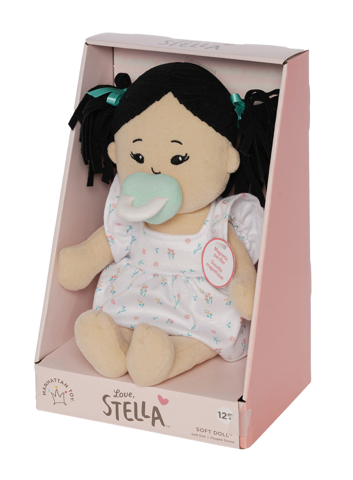 Soft doll with black hair and light beige skin wearing a dress with a light green magnetic pacifier on the doll's face while seated in pink packaged box at a quarter angle turn.