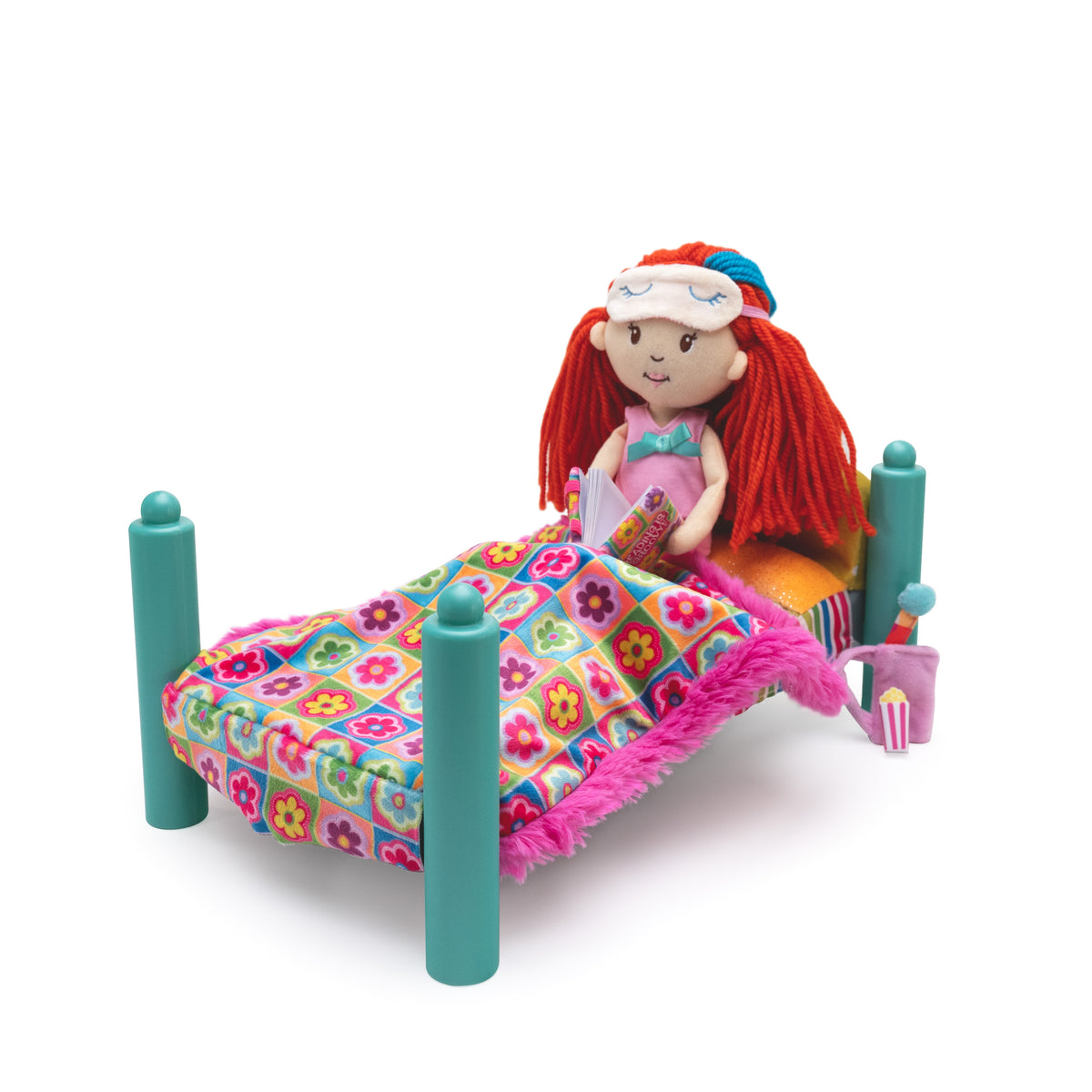 Doll sits on a miniature doll bed with teal posts and a colorful floral blanket covered in pink fringe, shown on a white background.