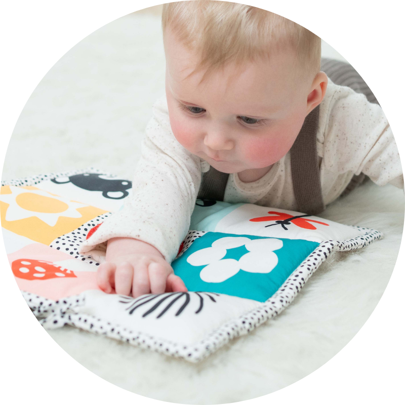 Infant baby boy on tummy laying on a colorful play mat reaching out his arm and gazing at the graphics on the mat while in a living room scene.