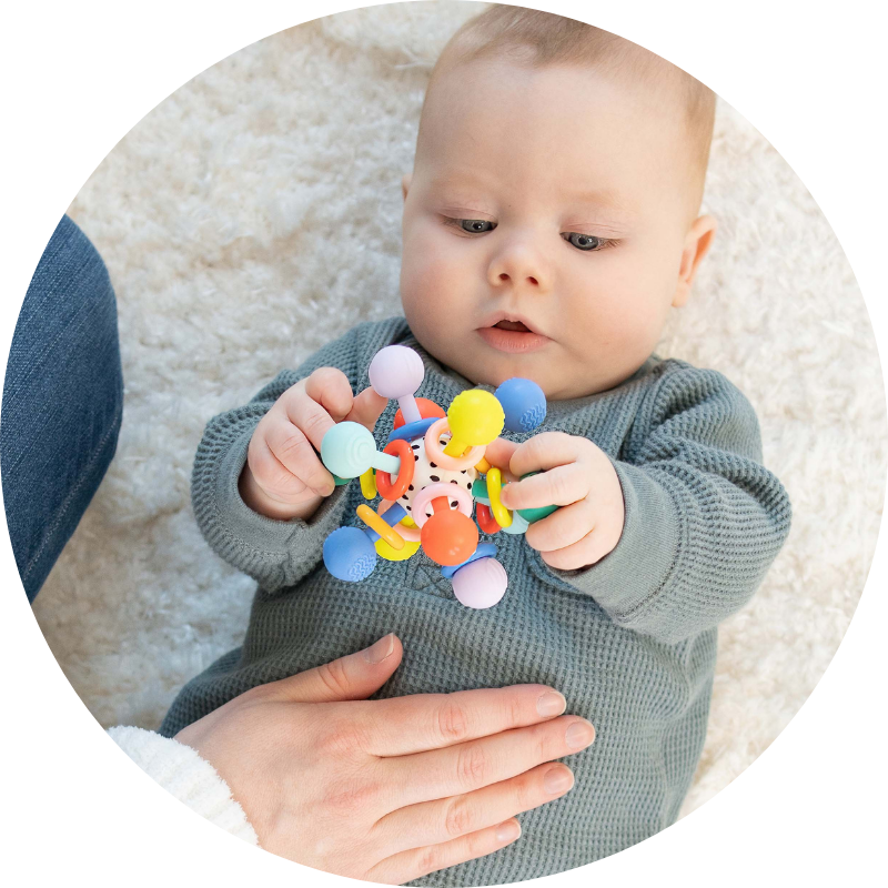Baby boy lying on back on plush cream rug holding onto a colorful teething rattle while gazing at it with mom kneeling nearby with her hand placed on his tummy.