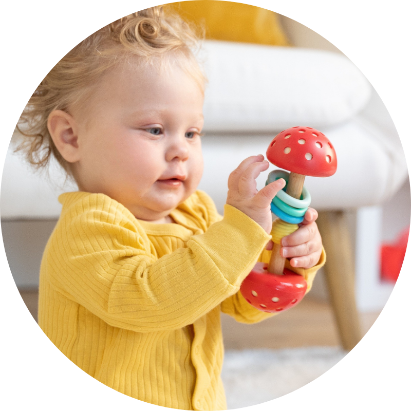 Baby girl seated on the floor in a living room setting holding onto a red colored and natural wood barbell rattle. Girl is gazing at the rattle in delight and wonder.