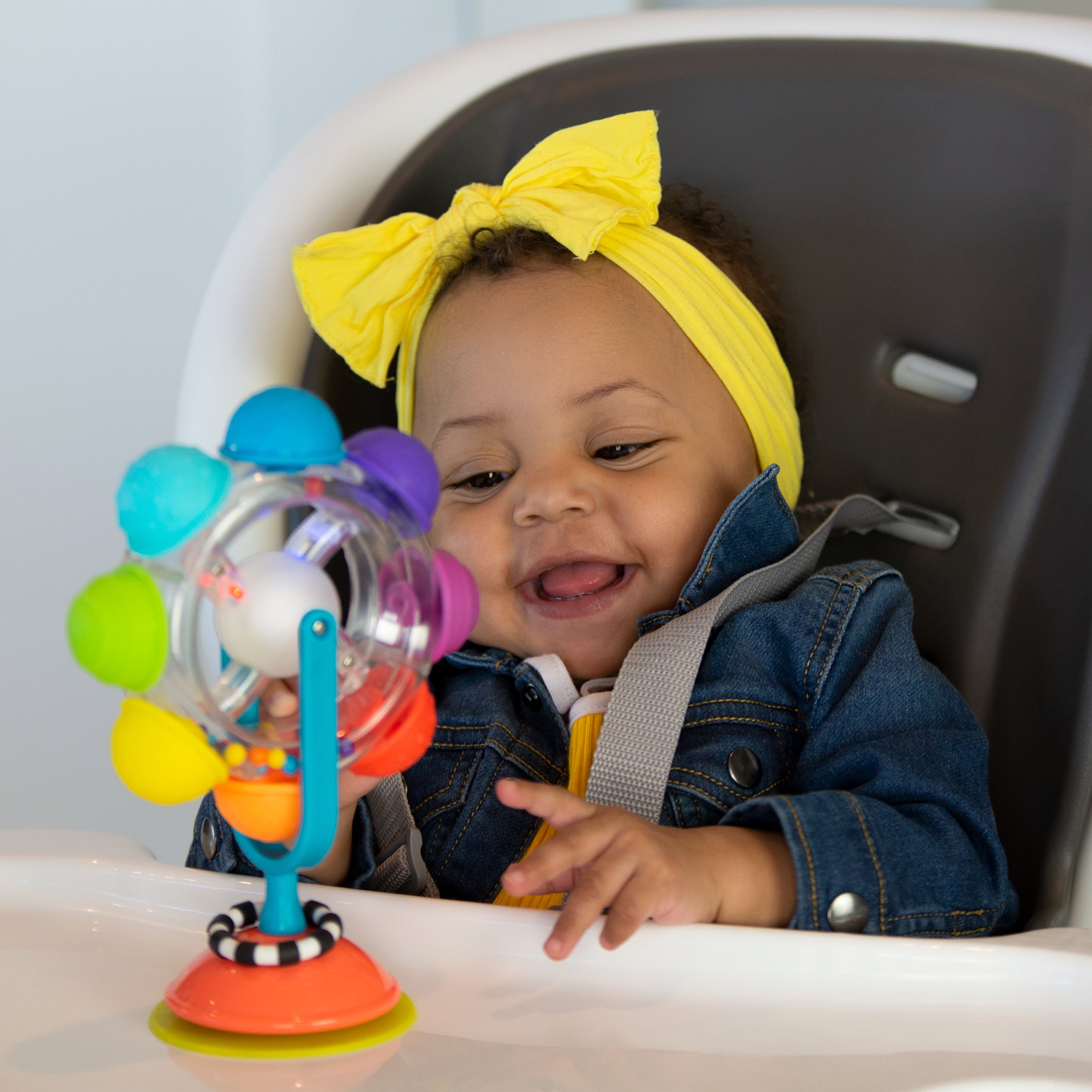 Child in a high chair with a colorful toy, wearing a yellow headband.