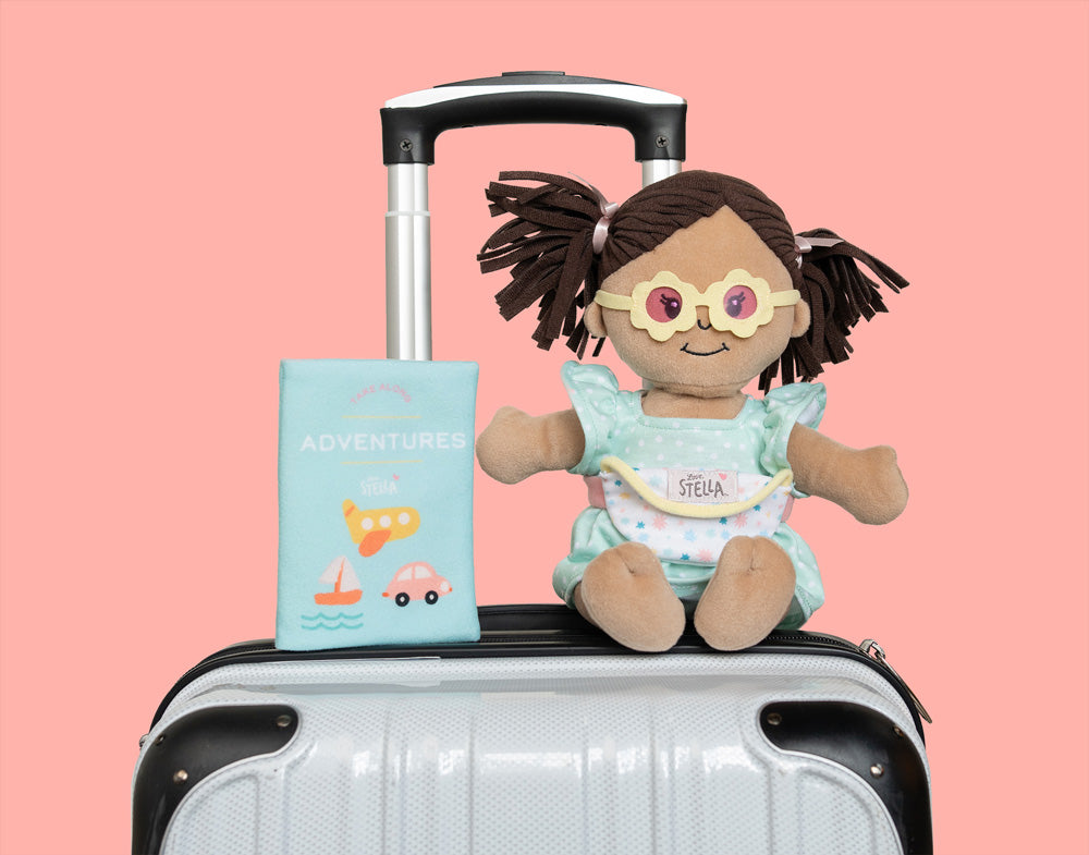Love, Stella adventure doll and travel book resting on a white suitcase against a pink background.