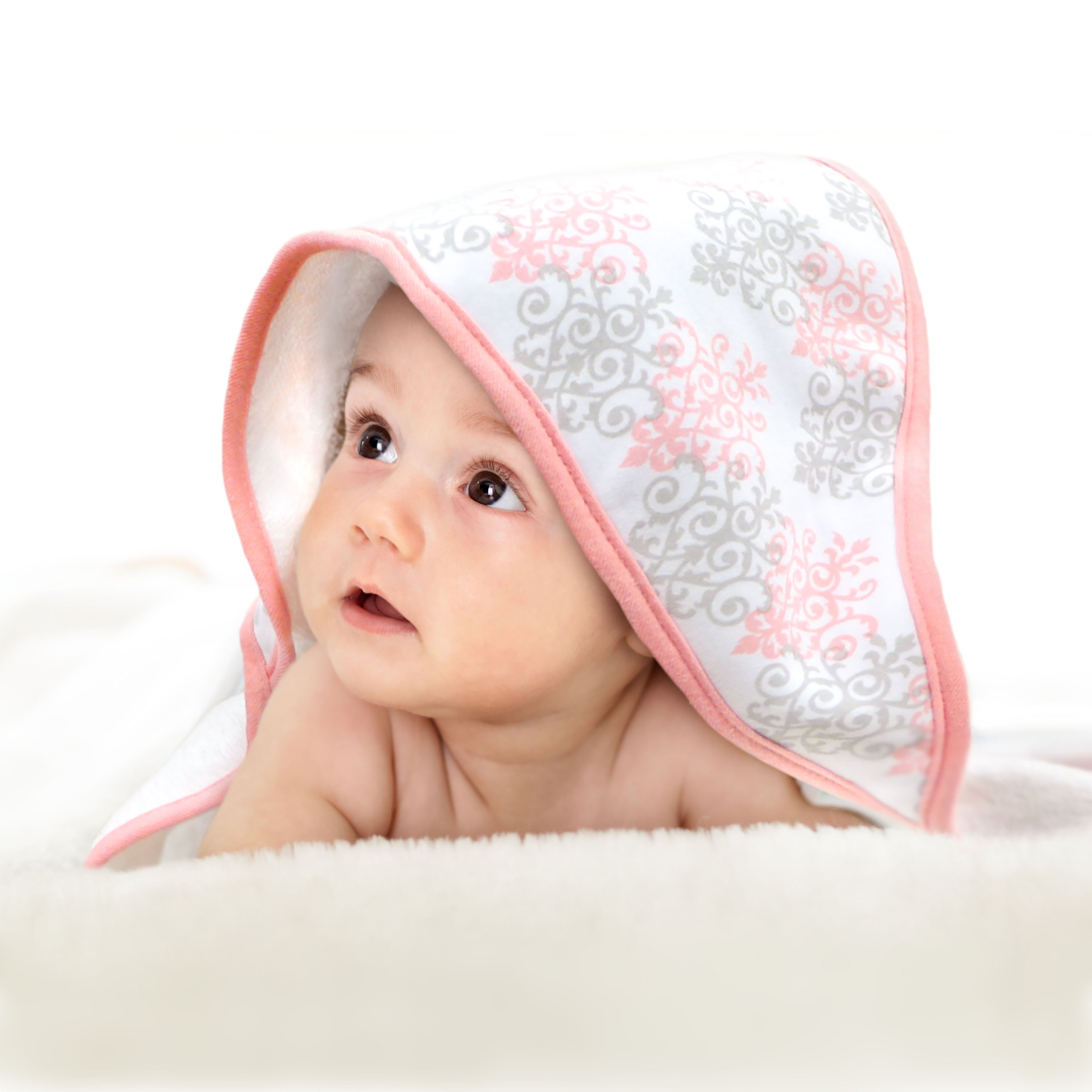 Adorable baby lying on a soft blanket with a pink and gray patterned hooded towel draped over their head, gazing upward.
