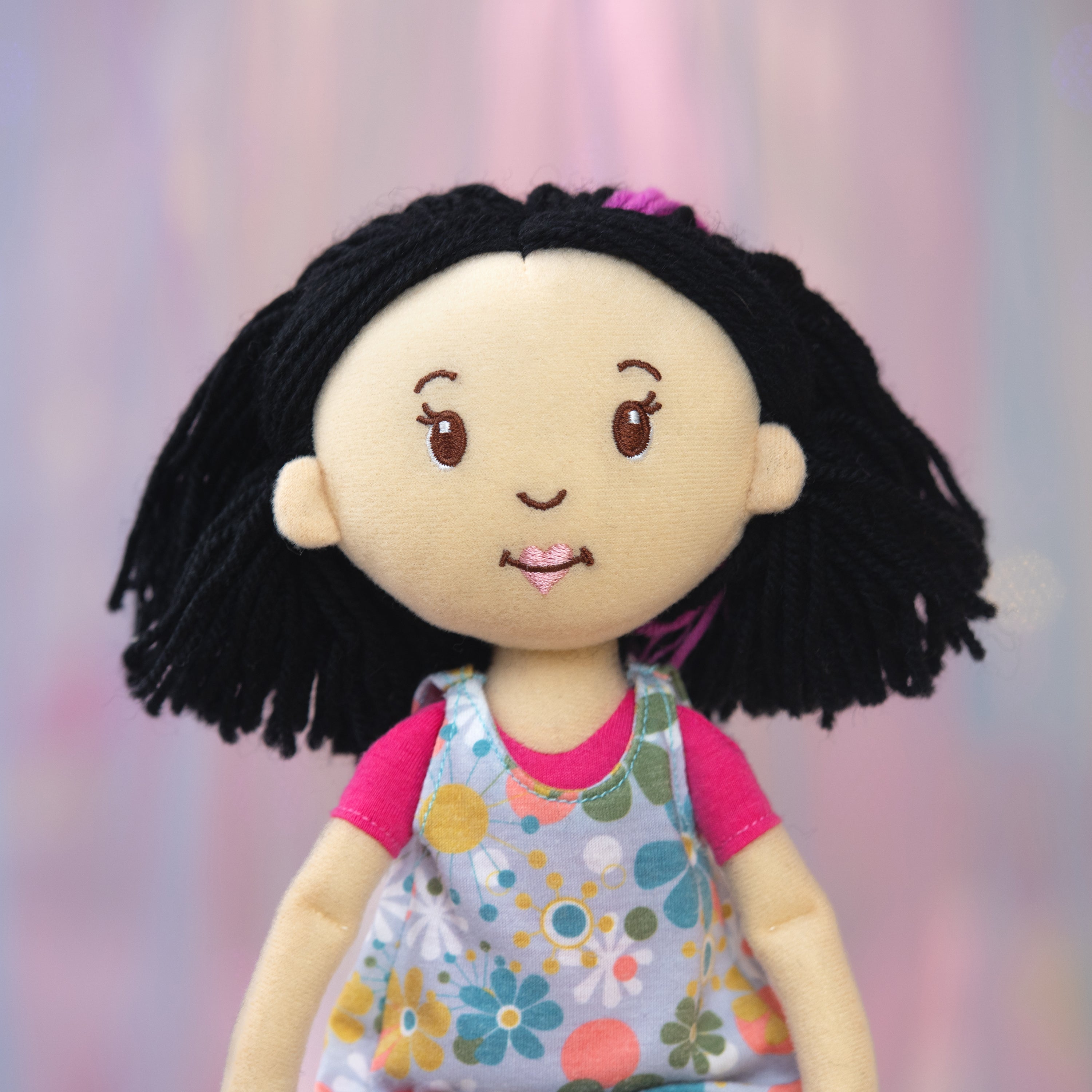Plush doll with black hair, pink shirt, and floral dress against a soft pink background