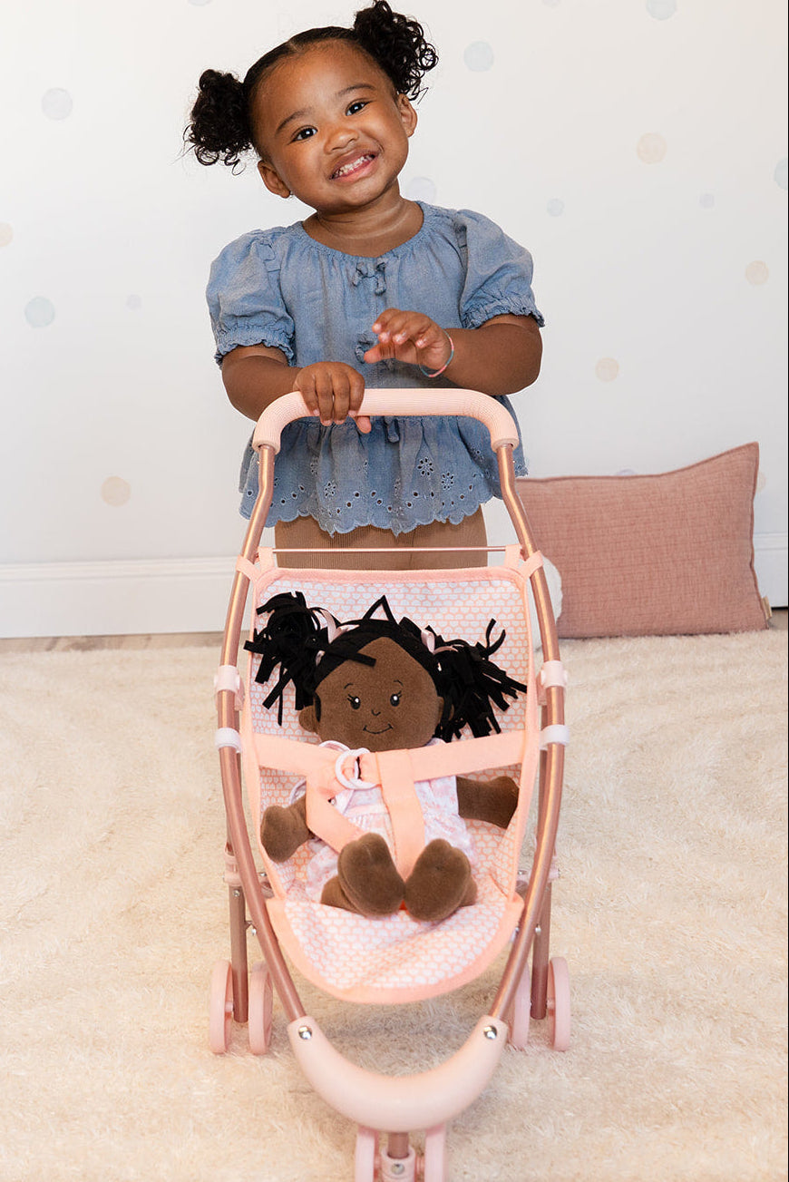 A little girl smiles wide while holding onto a pink doll stroller with a doll sitting inside.