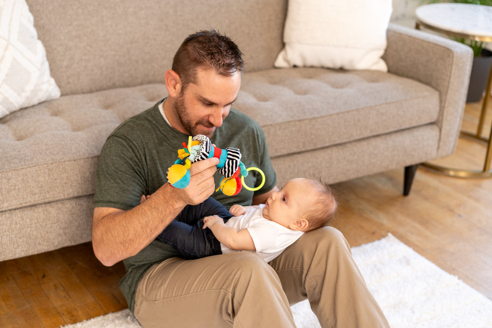 Father sitting on the floor with his baby on his lap, engaging them with a colorful sensory toy featuring various textures and patterns.