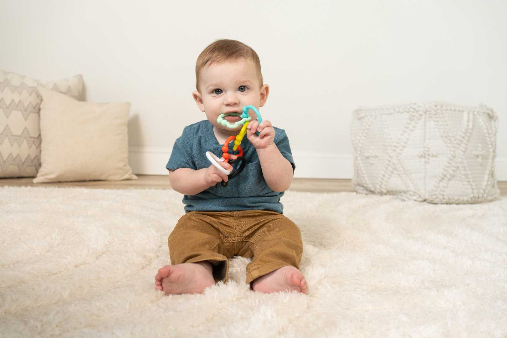 A baby sits on a rug holding multicolored bear-shaped rings in his mouth.