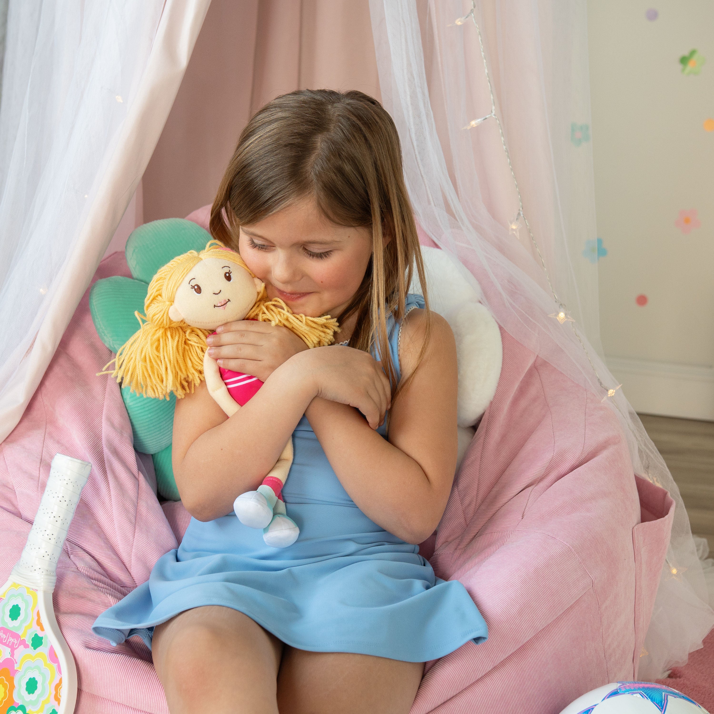 Young girl holding a plush toy in a cozy room with pink bean bag and decorative lights.