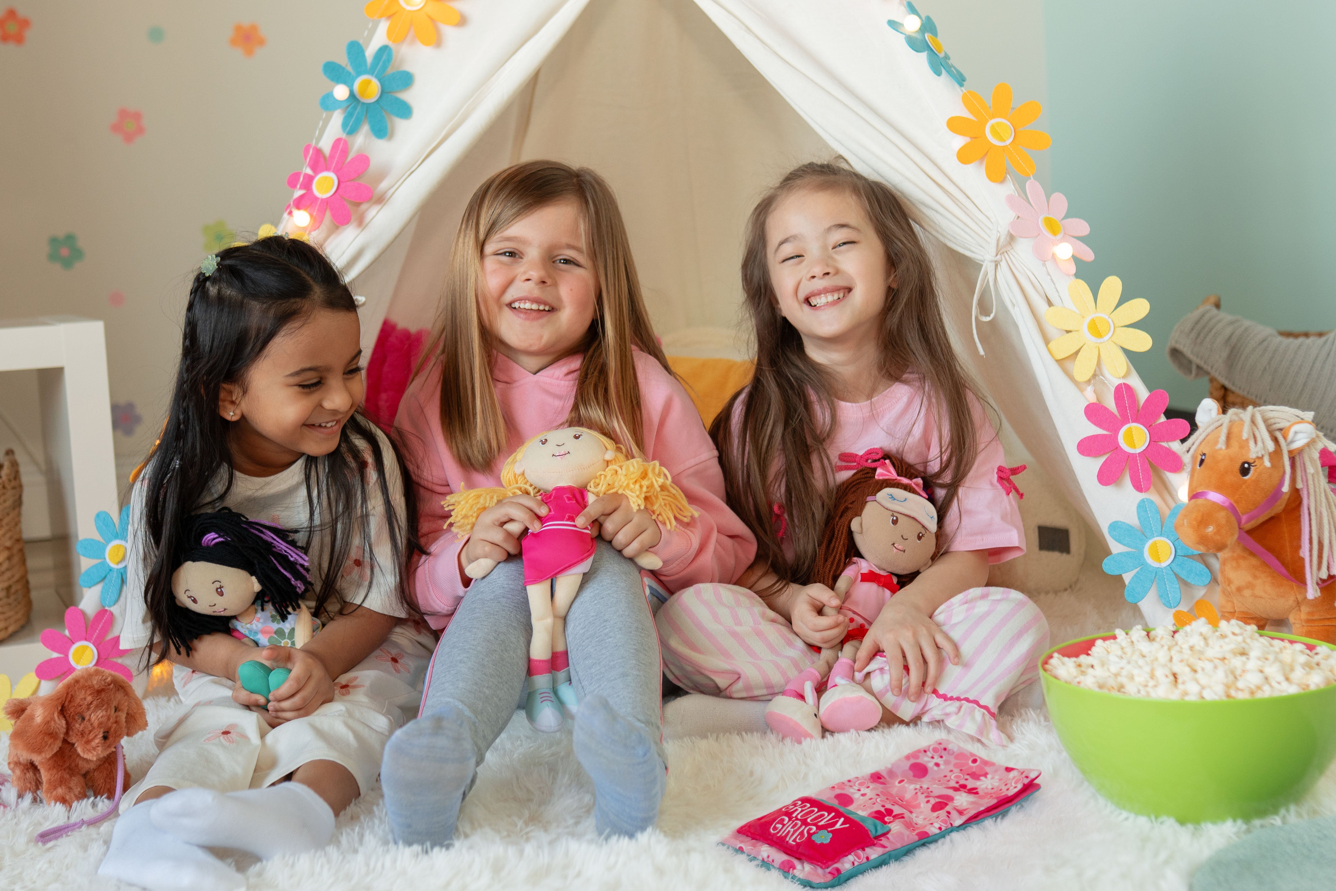 Three children sitting inside a play tent with toys and popcorn, surrounded by colorful flowers.