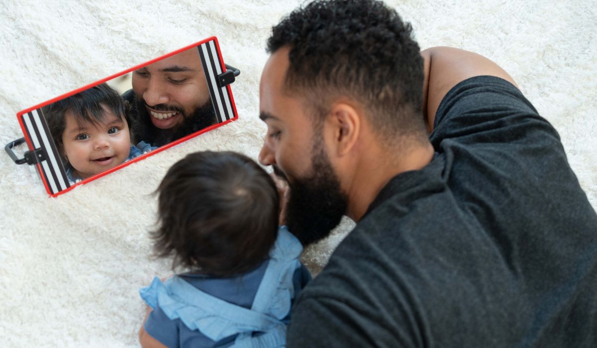 Father and baby girl lying on their tummies smiling together while looking into a sensory tummy time mirror with black and white stripes and a red frame.