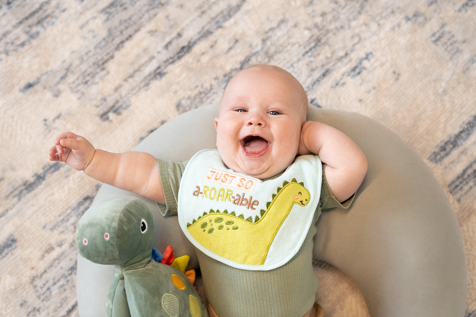 A grinning baby laying down on a pillow with a green plush dinosaur next to him.