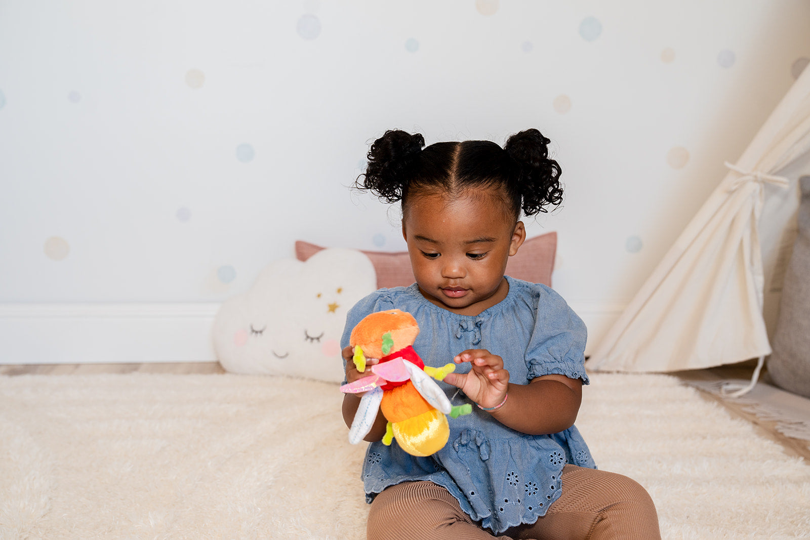 A toddler looks down at the multicolored plush dragonfly puppet in her hands.