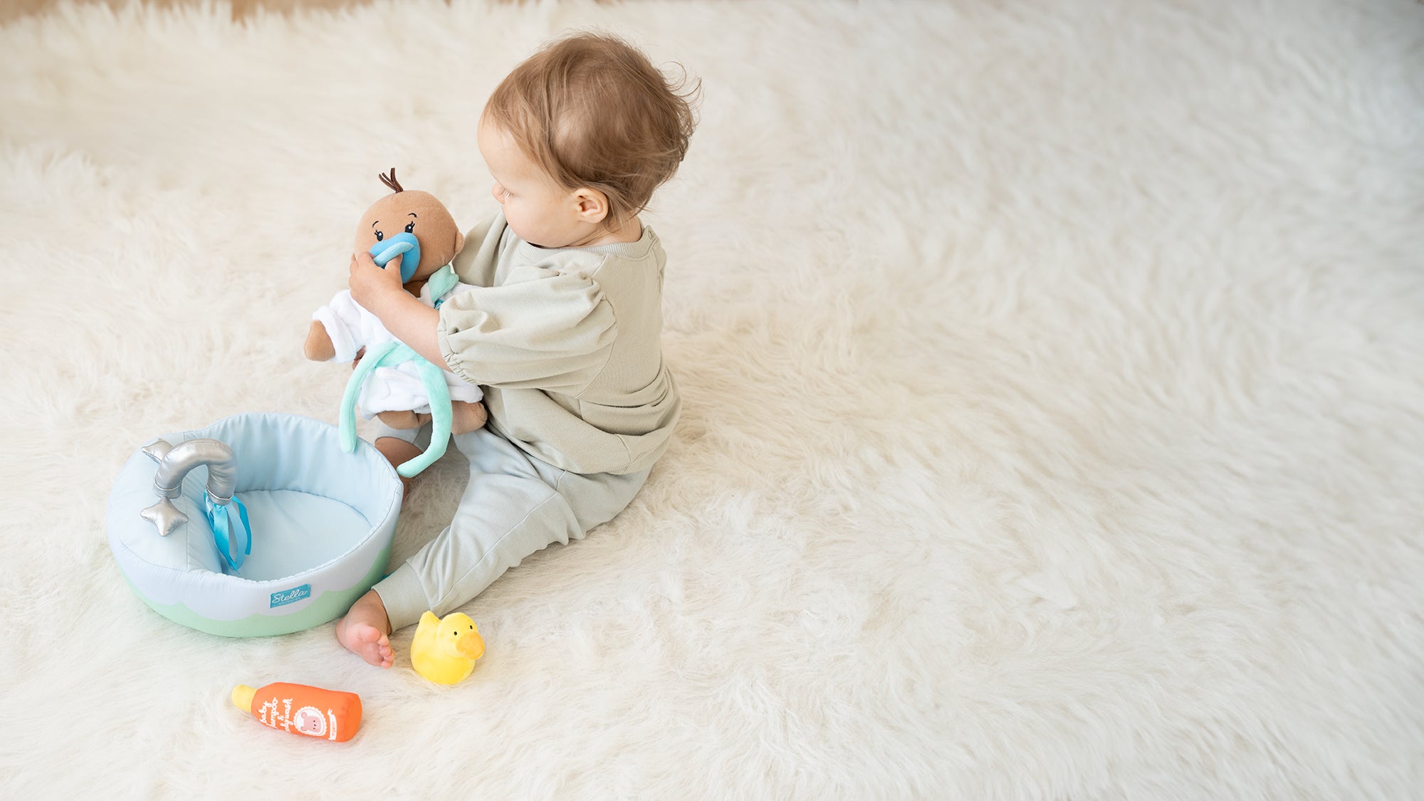 Child on a fluffy white rug holding a doll, with a small toy bathtub, duck, and bottle nearby.