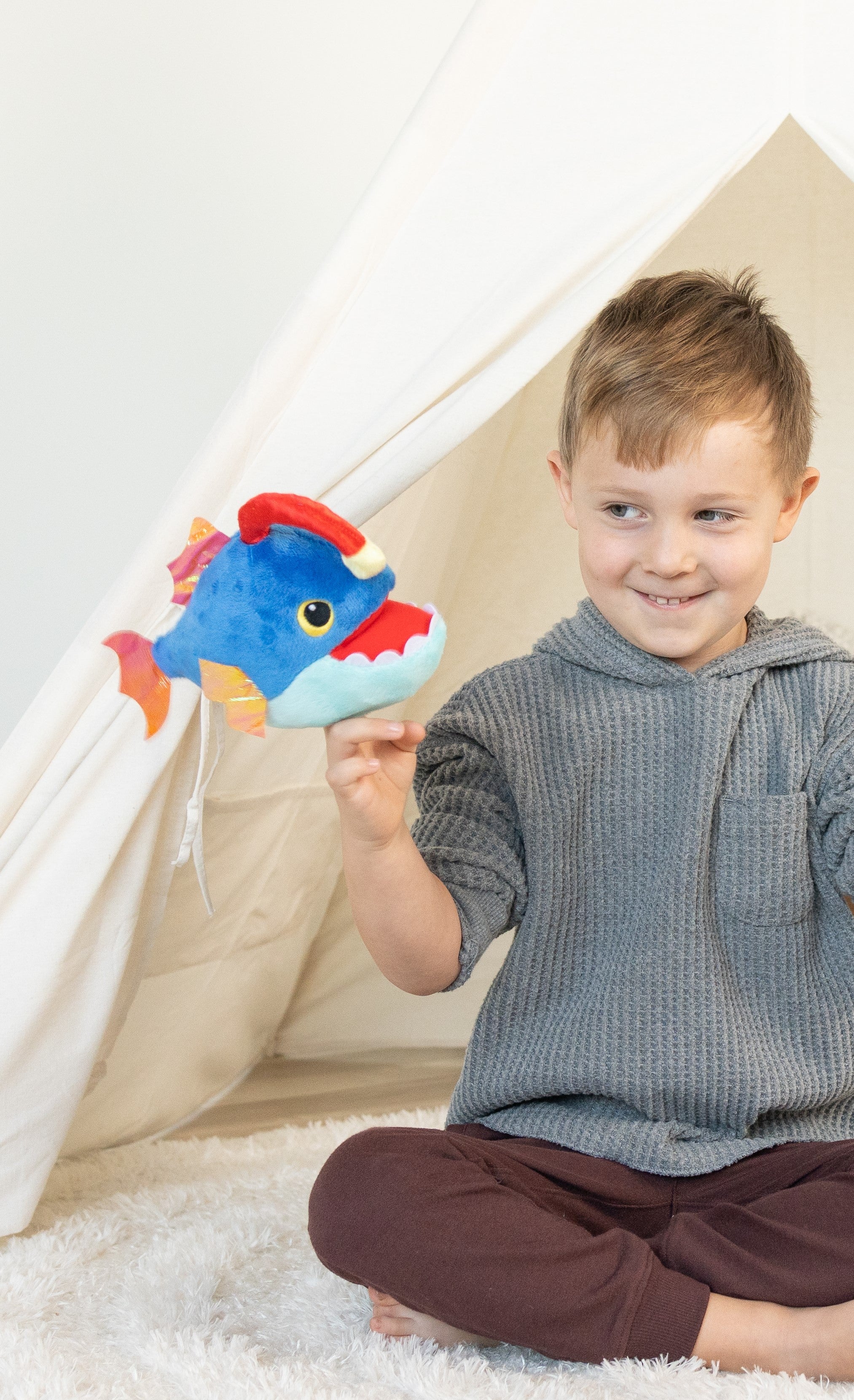 A young boy sits cross-legged on the floor with a blue plush anglerfish finger puppet on his hand.