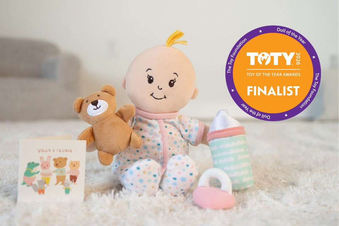 Soft baby doll seated on rug with teddy bear, bottle, mini paper book and pacifier next to a TOTY award Finalist seal on right side.