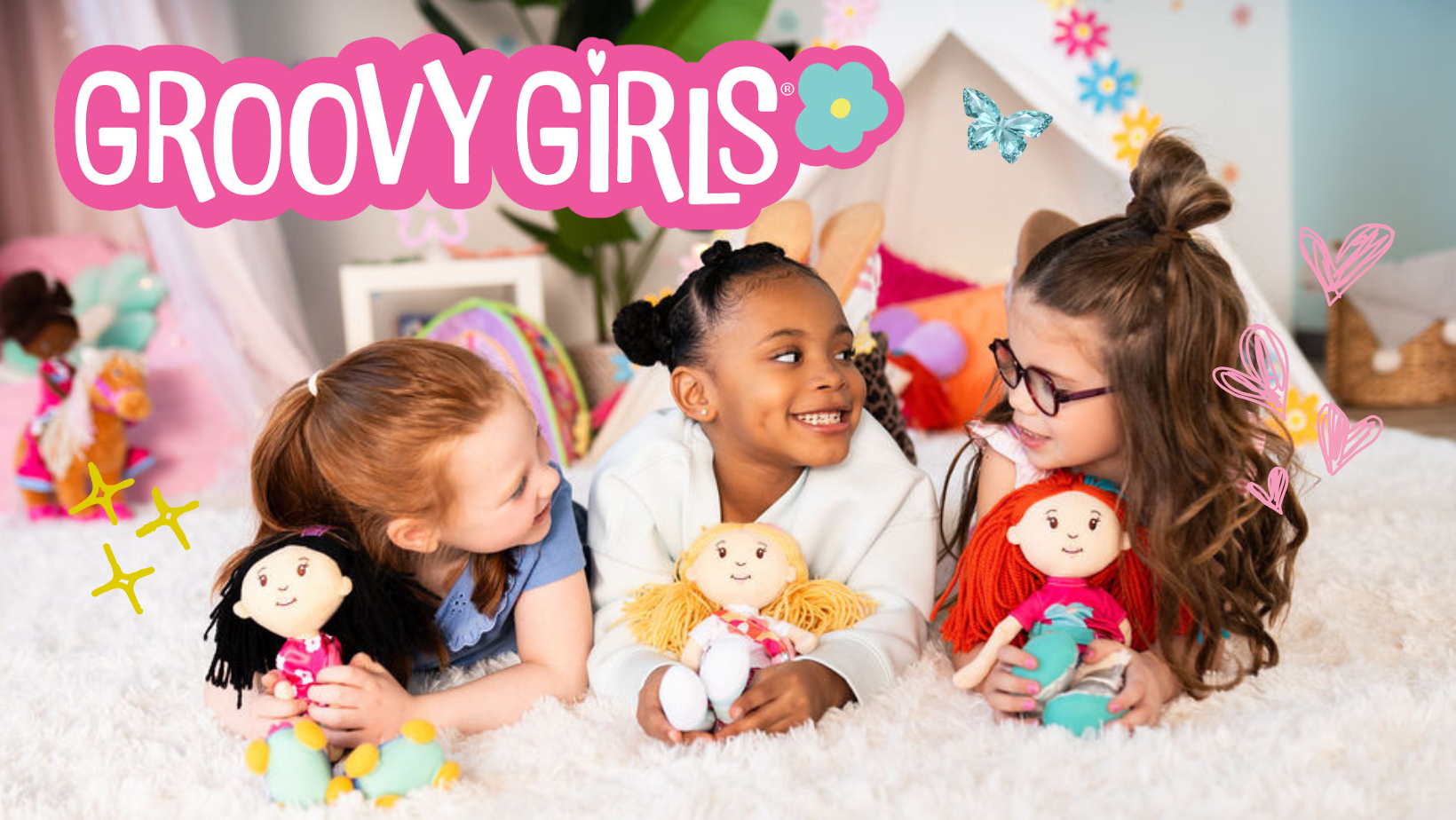 three 1st grader girls laying on on their tummies shoulder to shoulder holding soft dolls in front of them while smiling at one another in a girls room scene with Groovy Girls logo overlayed above them.