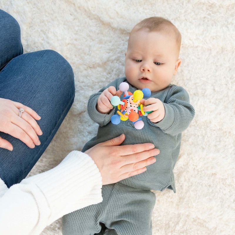 Infant baby boy laying on back on a cream rug holding a colorful grasping toy over his chest while looking at the toy with his mom kneeling next to him with her hand on his belly.