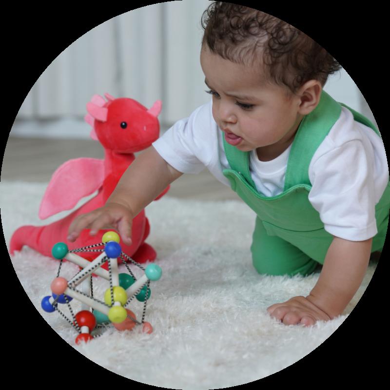 Baby boy in crawling position on a cream rug, reaching for wooden baby rattle with a bright red dragon in the background.