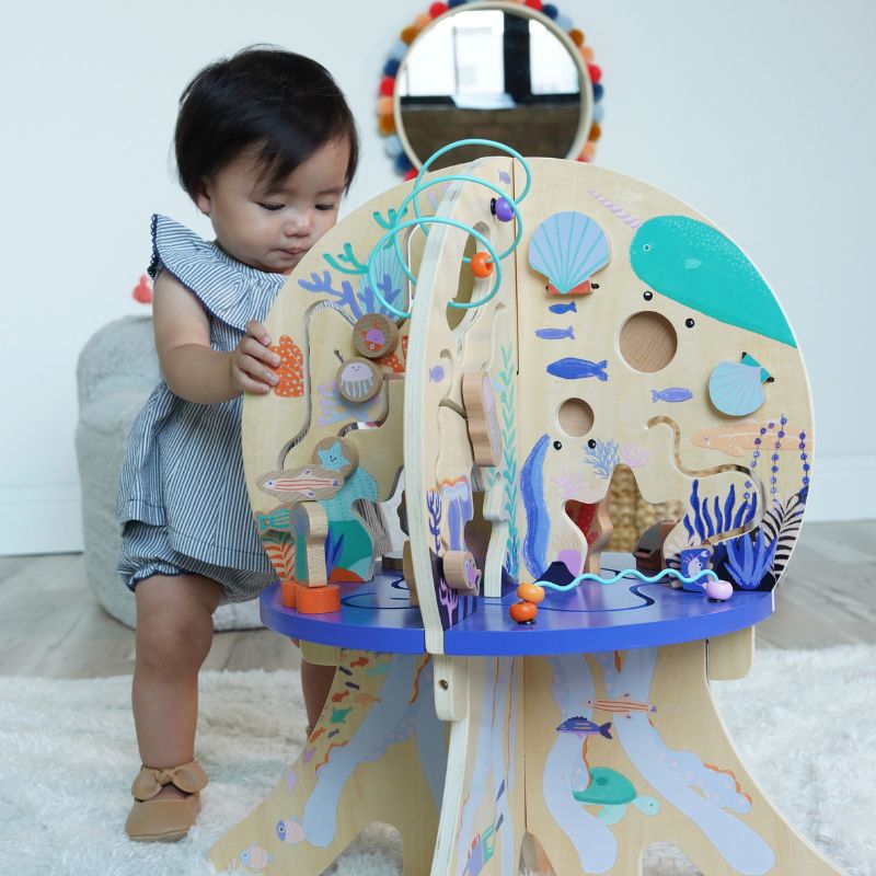 12 month old girl standing while holding onto and playing with a large wood activity center with under water sea themed graphics in a living room setting 