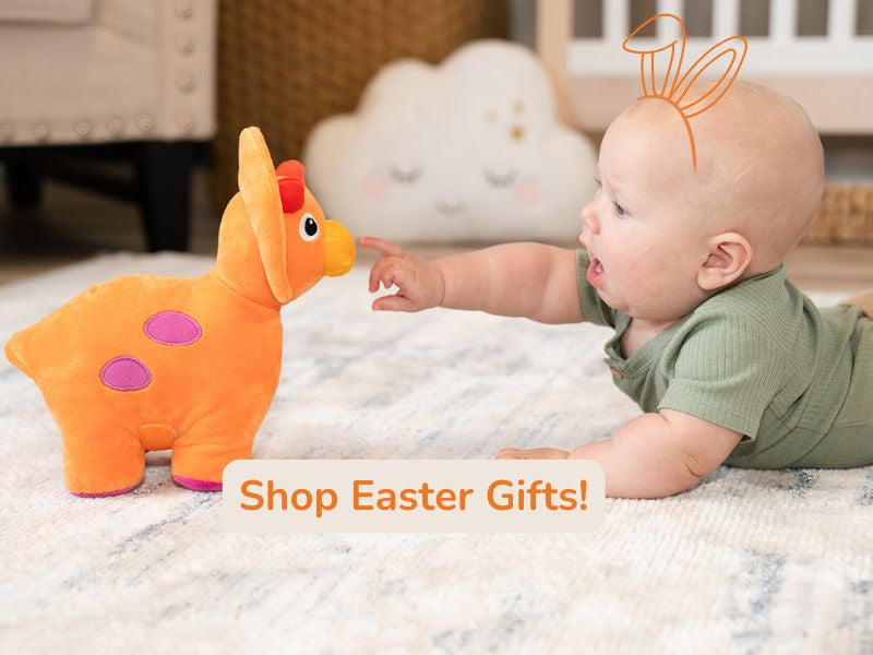 Baby boy laying on tummy with hand-drawn orange bunny ears on head, reaching towards orange pluch dinosaur in a nursery room scene. Overlay text reads Shop Easter Gifts!