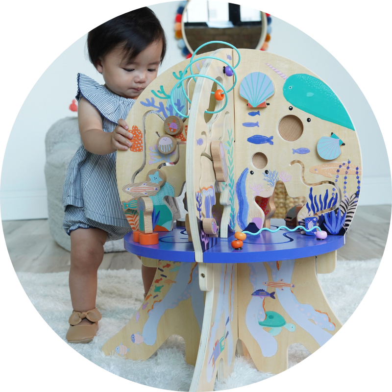 Toddler age girl standing slightly behind and next to a large wood activity center toy with underwater sea themed graphics. Girl is holding onto the activity center as she gazes at the toy's features while standing in a living room setting with a small mirror hanging behind her on the wall. 