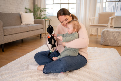 Smiling mother sitting on a rug holding her baby, who is reaching out toward a black and white sensory bear lovey toy.
