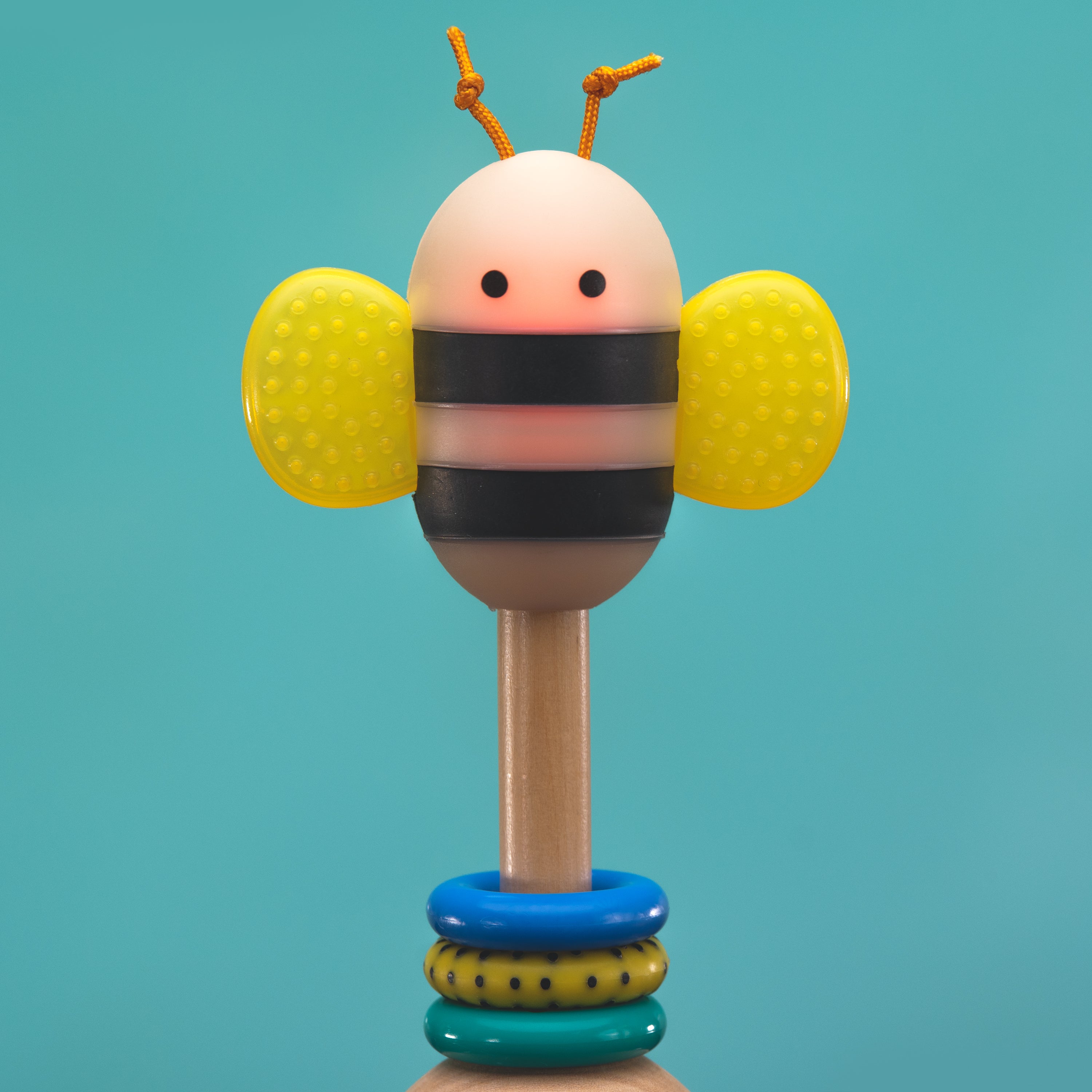 Bee themed baby rattle with glowing body, yellow textured wings, and blue, yellow, and green rings against a teal background.