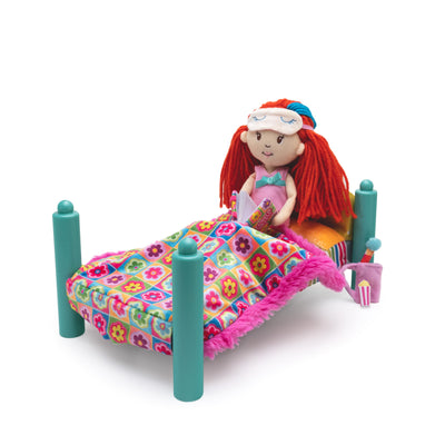 Doll sits on a miniature doll bed with teal posts and a colorful floral blanket covered in pink fringe, shown on a white background.