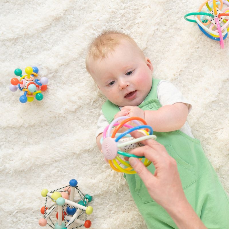 Baby boy laying on his back on a plush cream rug with colorful teether/rattle toys scattered around him as he gazes at a teether toy being held over his tummy by his mom. 