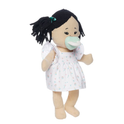 Soft doll with black hair and light beige skin wearing a dress with a light green magnetic pacifier on the doll's face while standing.