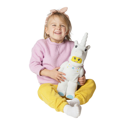 Smiling young girl in a pink sweater holding a plush LEGO-style unicorn toy while sitting cross-legged.