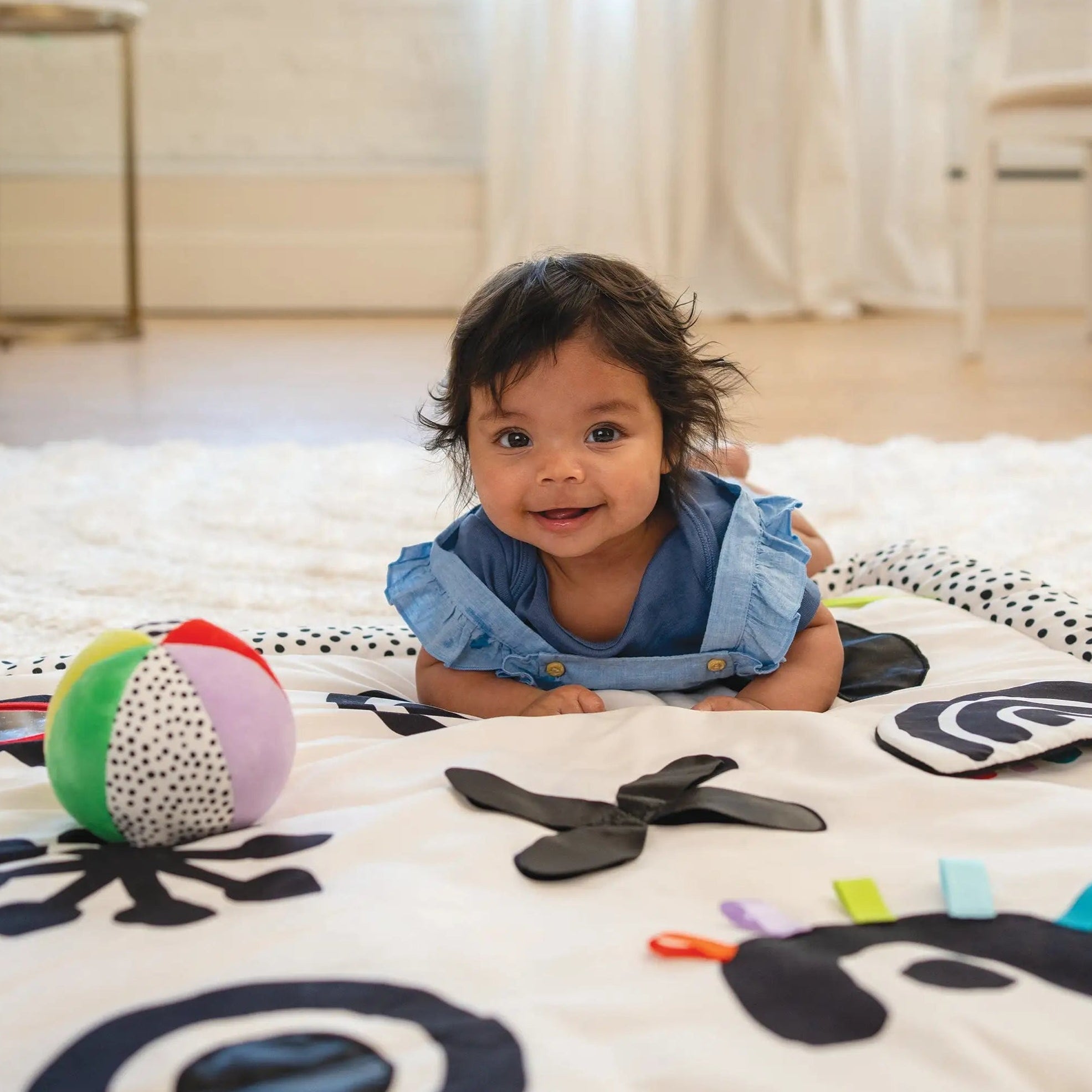 Smiling baby in a blue outfit during tummy time on a high-contrast developmental play mat with a colorful ball and textured fabric activities.