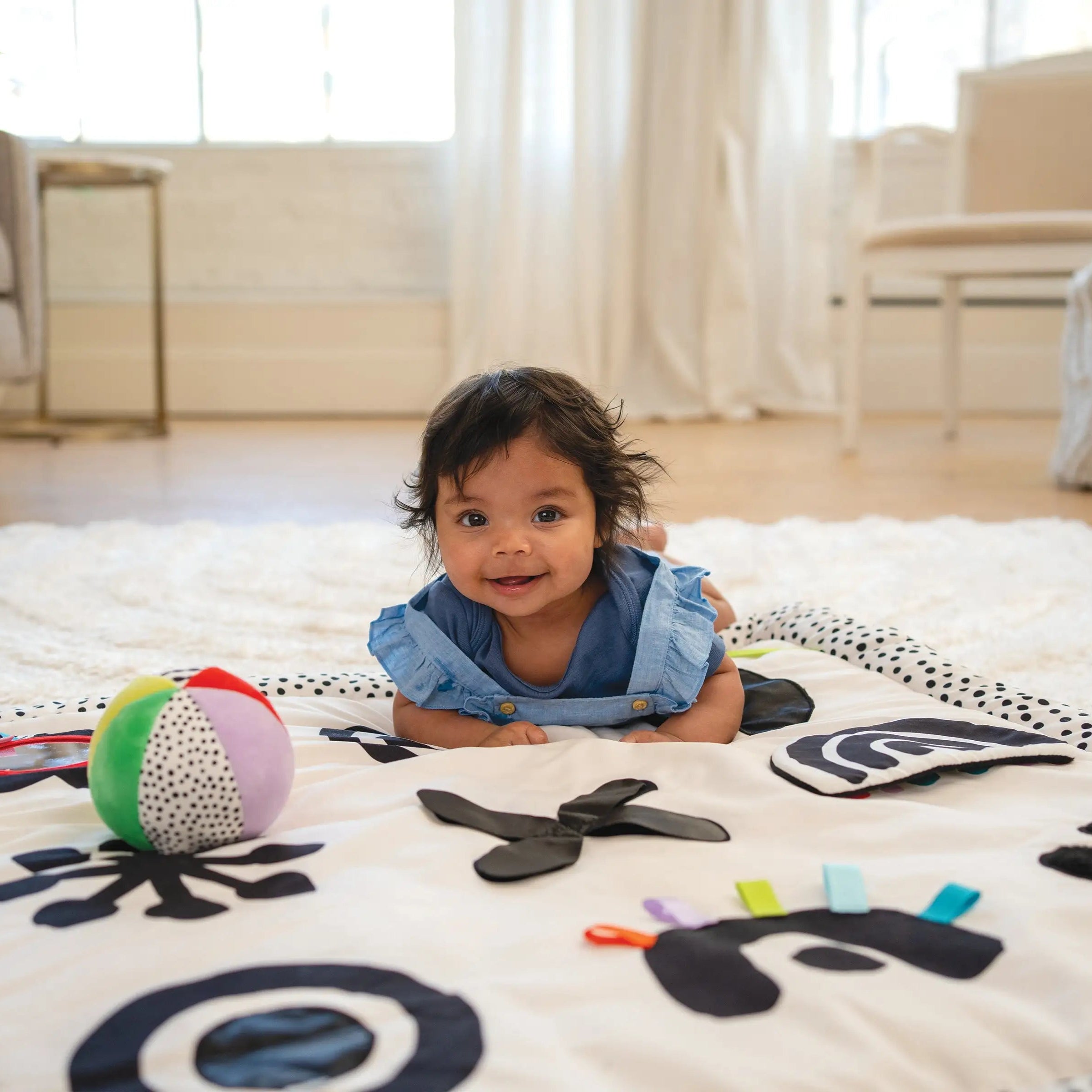 Smiling baby in a blue outfit during tummy time on a high-contrast developmental play mat with a colorful ball and textured fabric activities.