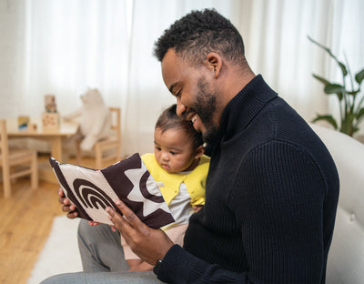 Dad and baby seated on couch looking at soft mirror booth while smiling.