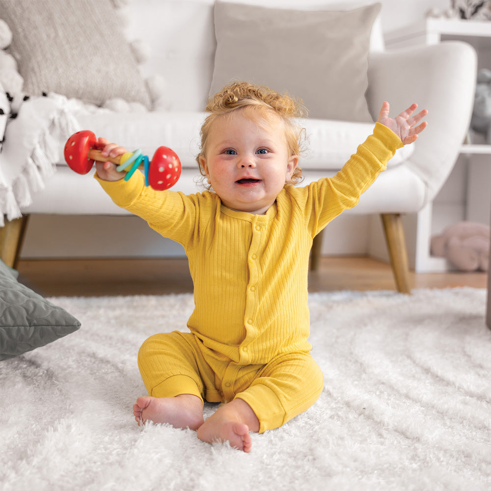 Baby in a yellow onesie sitting on a fluffy rug, holding a colorful rattle, with a couch and pillows in the background.