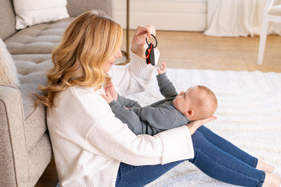 Mother sitting on the floor holding her baby while showing a black and red toy; baby reaches out with curiosity and focus.