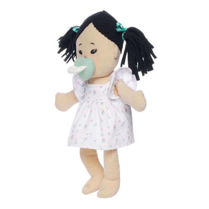 Soft doll with black hair and light beige skin wearing a dress with a light green magnetic pacifier on the doll's face while standing.