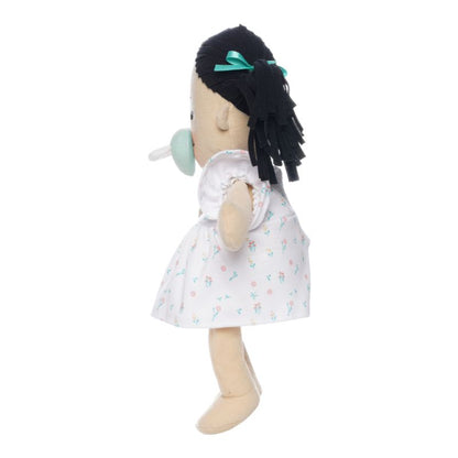 Soft doll with black hair and light beige skin wearing a dress with a light green magnetic pacifier on the doll's face while standing.