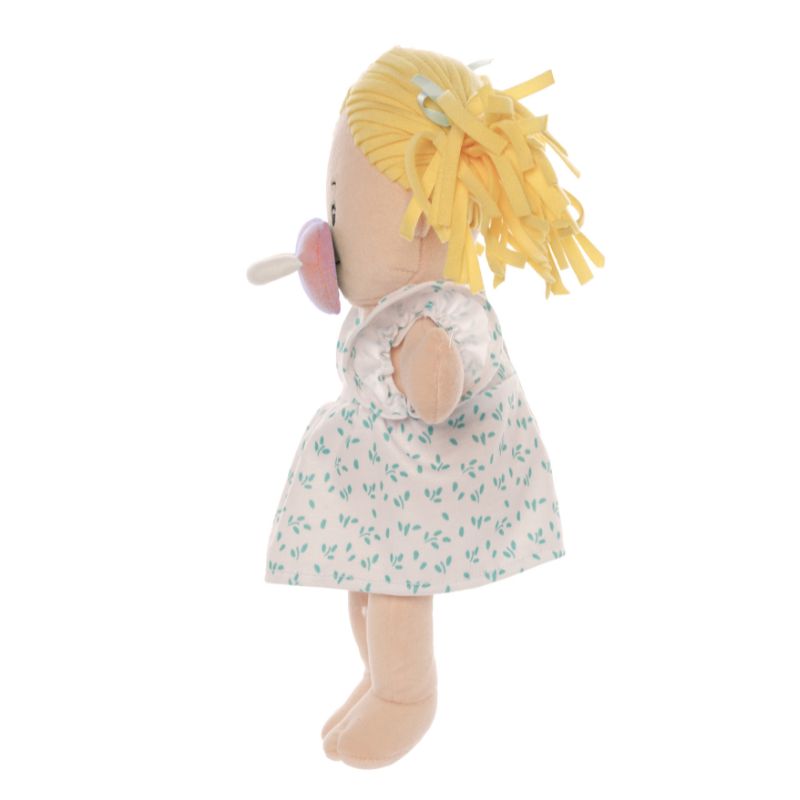 Peach skinned soft doll in a standing 
 side profile position with blonde hair pigtails wearing a dress with a pink pacifier on the face of the doll. 