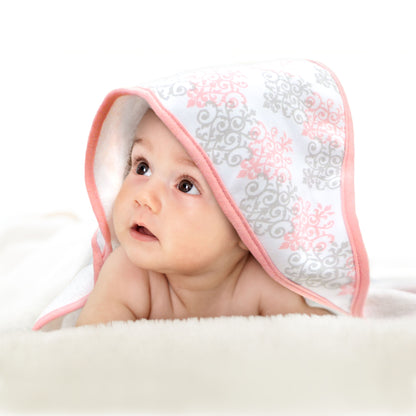 Adorable baby lying on a soft blanket with a pink and gray patterned hooded towel draped over their head, gazing upward.