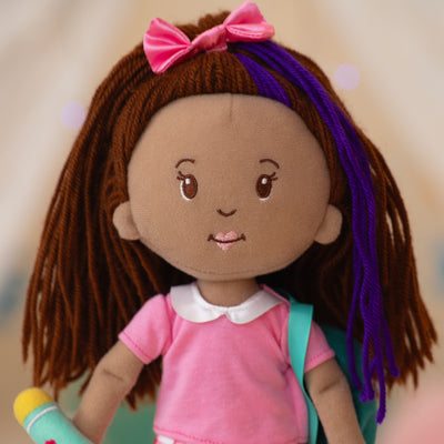 close up of a soft doll wearing a pink collared shirt, holding a teal backpack and teal water bottle