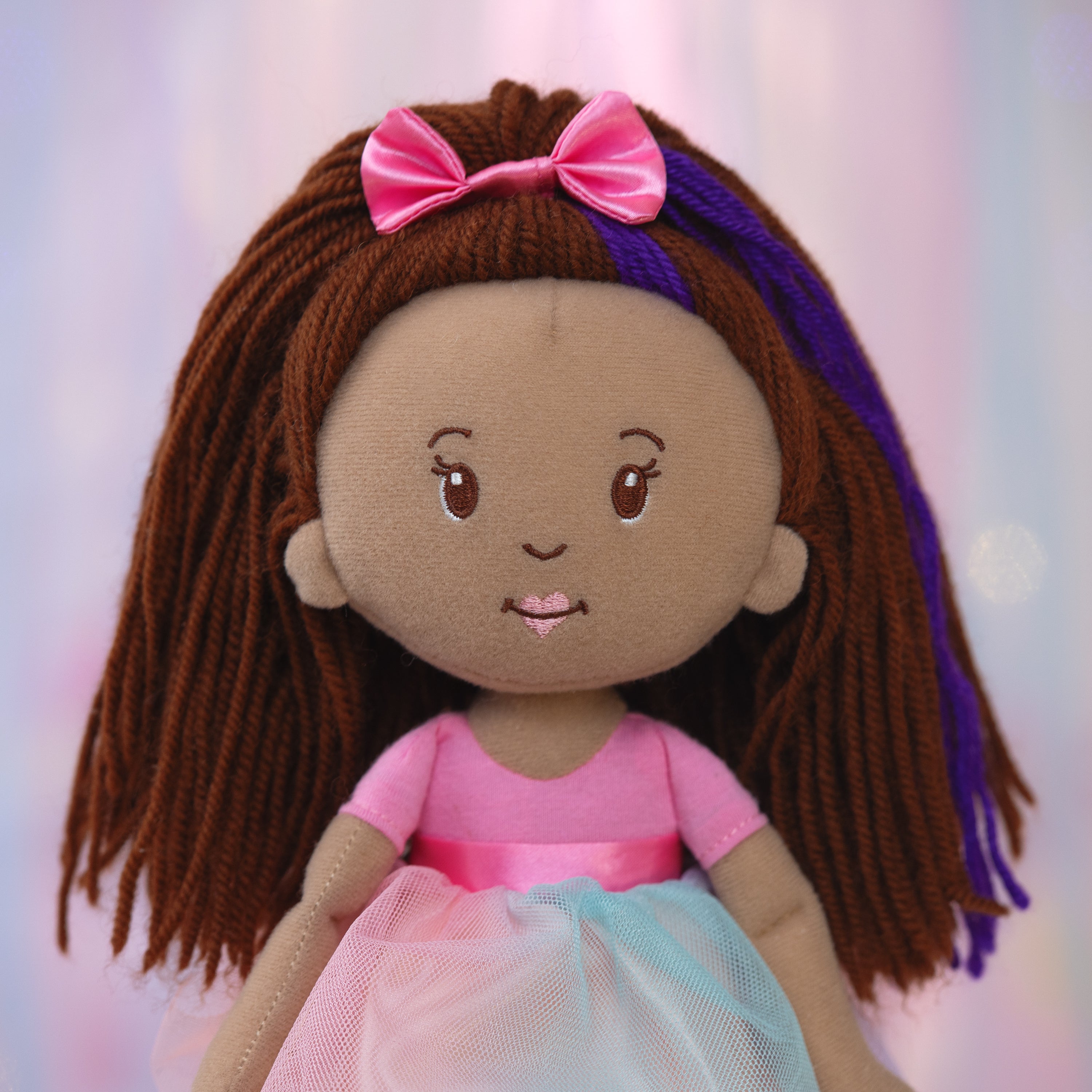 Plush doll with brown hair, pink bow, and pink dress on a pink and purple background