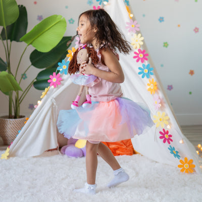 Child in a colorful dress holding a doll inside a decorated play area with flowers and a teepee.