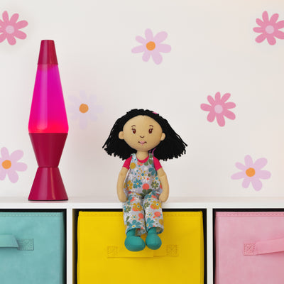 Doll with floral overalls sitting on a shelf with a pink lava lamp and colorful boxes against a wall with flower decals.
